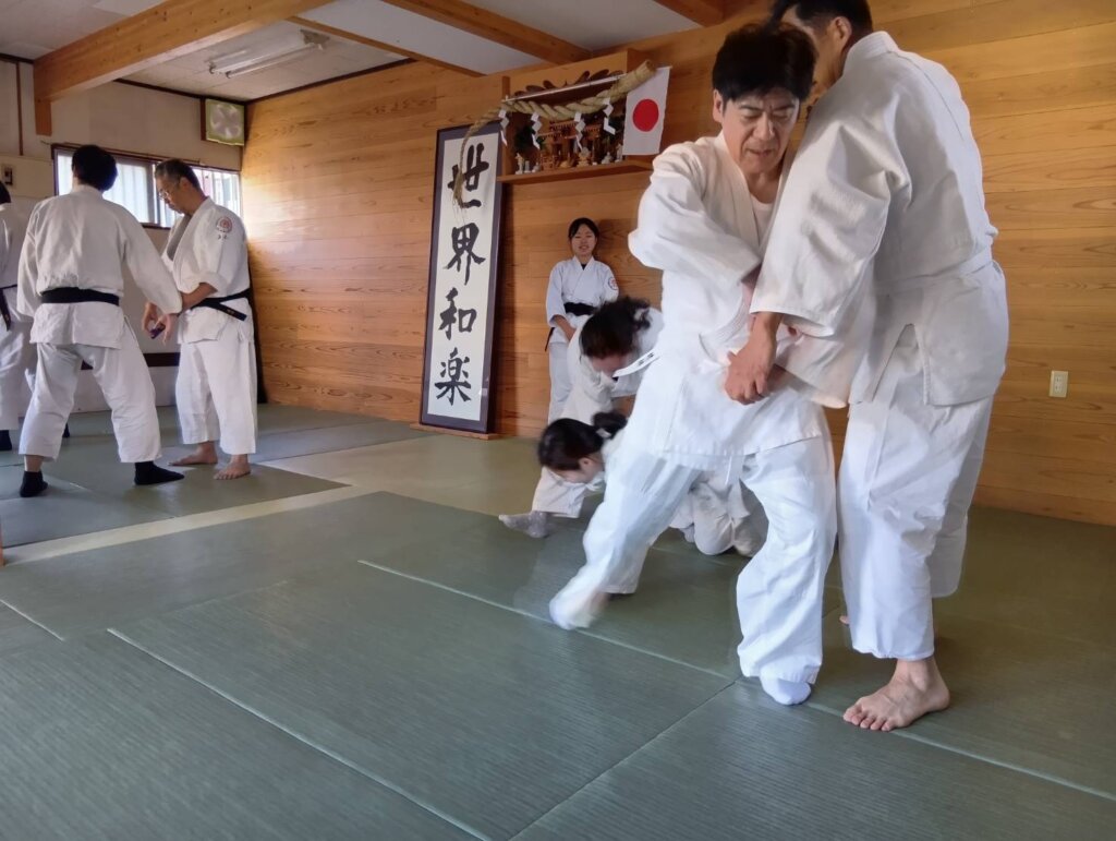 木造の道場で、複数の門人が白い稽古着で技の稽古を行っている。背景には「世界和楽」と書かれた大きな掛け軸が掲げられている。
