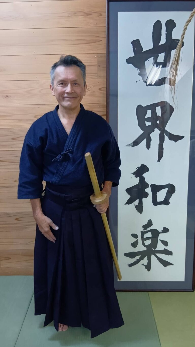 木刀を手にした男性が道場で立っている。背後には「世界和楽」と書かれた書が掛けられている。